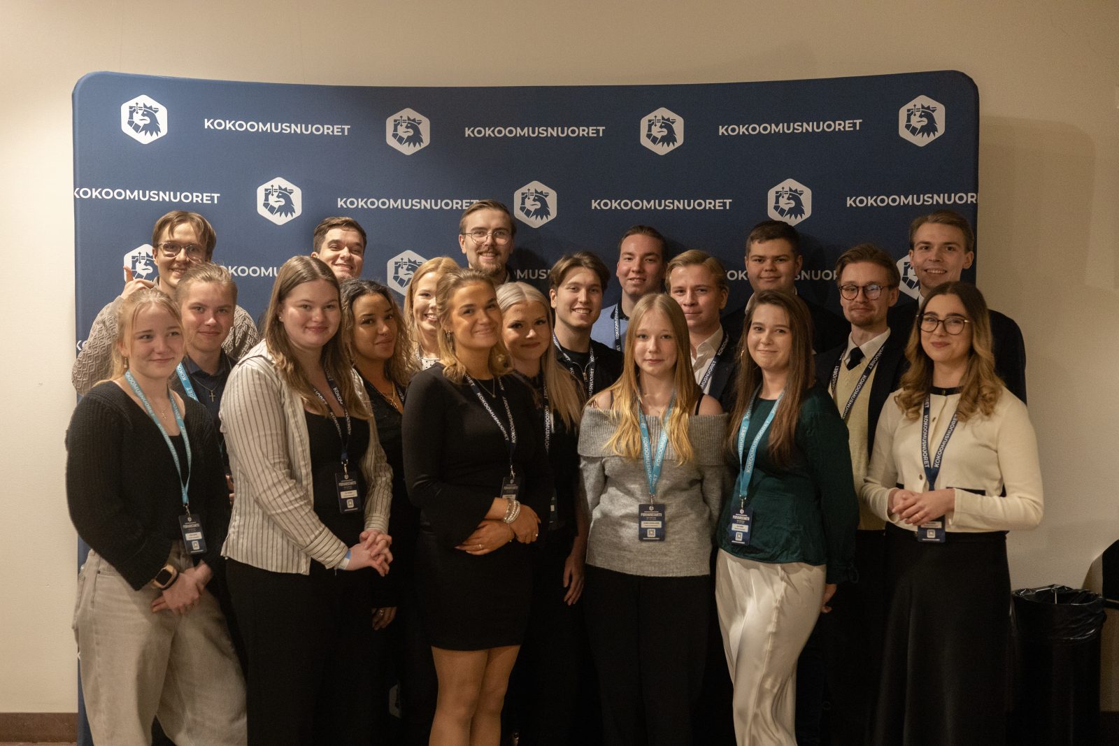 Suuri ryhmä nuoria aikuisia poseeraa sisätiloissa Kokoomusnuorten sinistä bränditaustaseinää vasten. Henkilöt seisovat tiiviissä ryhmässä useassa rivissä, suurin osa hymyilee ja kaikilla on tapahtumapassit kaulassaan. Taustaseinässä näkyy Kokoomusnuorten nimi ja leijonalogo.