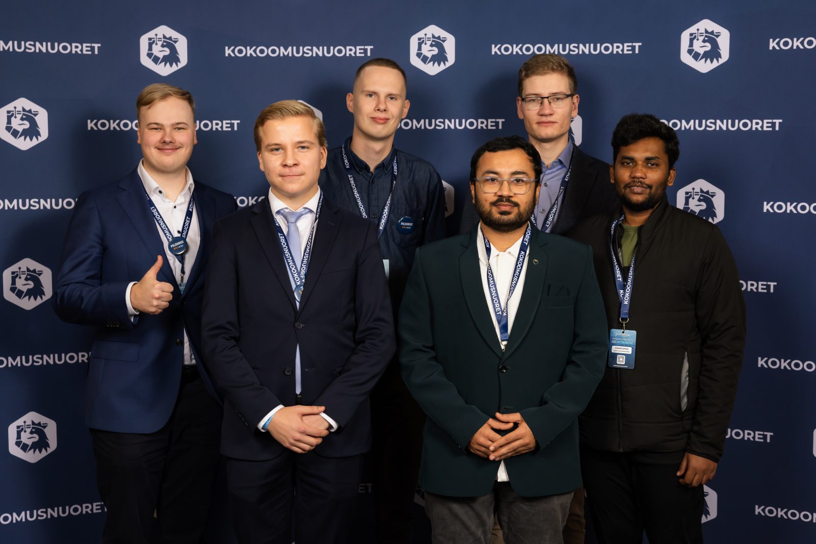 Ryhmäkuva kuudesta henkilöstä Kokoomusnuorten brändätyn taustaseinän edessä. Kaikki seisovat rivissä ja katsovat kameraan. Vasemmalla oleva henkilö näyttää peukkua, ja muut poseeraavat neutraalisti tai kevyesti hymyillen. Kaikilla on kaulanauhat ja kuvassa näkyy siistit, tapahtumaan sopivat vaatteet. Tausta on sinisävyinen ja siinä toistuu Kokoomusnuorten logo.