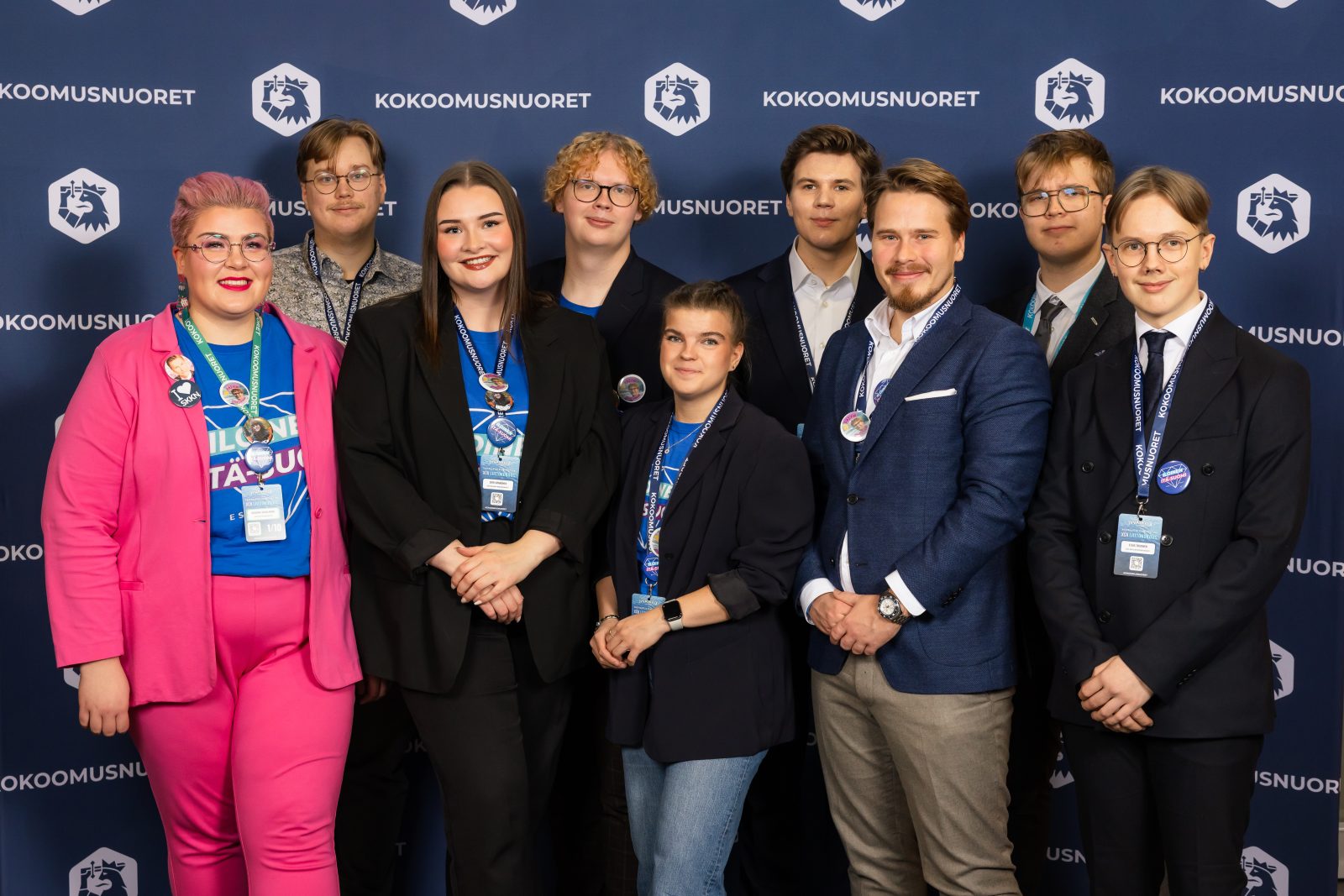 Ryhmäkuva, jossa yksitoista nuorta aikuista poseeraa Kokoomusnuorten kuvausseinän edessä. Eturivissä seisoo seitsemän henkilöä ja takarivissä neljä. Osalla heistä on kaulassaan liittokokouspassi ja sinisävyisiä kampanjapaitoja, ja yksi henkilö erottuu kirkkaan pinkillä puvulla. Kaikki katsovat kameraan ja hymyilevät.