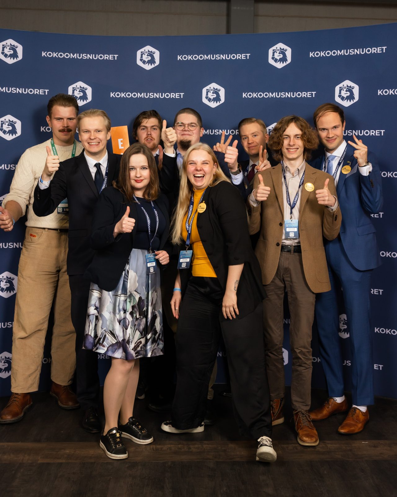 Ryhmäkuva, jossa yksitoista nuorta aikuista poseeraa Kokoomusnuorten kuvausseinän edessä. He ovat asettuneet kahteen riviin: eturivissä kuusi henkilöä ja takarivissä viisi. Useimmat näyttävät peukkua tai muuta iloista käsimerkkiä. Henkilöillä on tapahtumatunnisteet kaulassaan, ja heidän pukeutumisensa vaihtelee puvusta rennompaan juhlapukeutumiseen. Kaikki näyttävät hyväntuulisilta ja katsovat kohti kameraa.