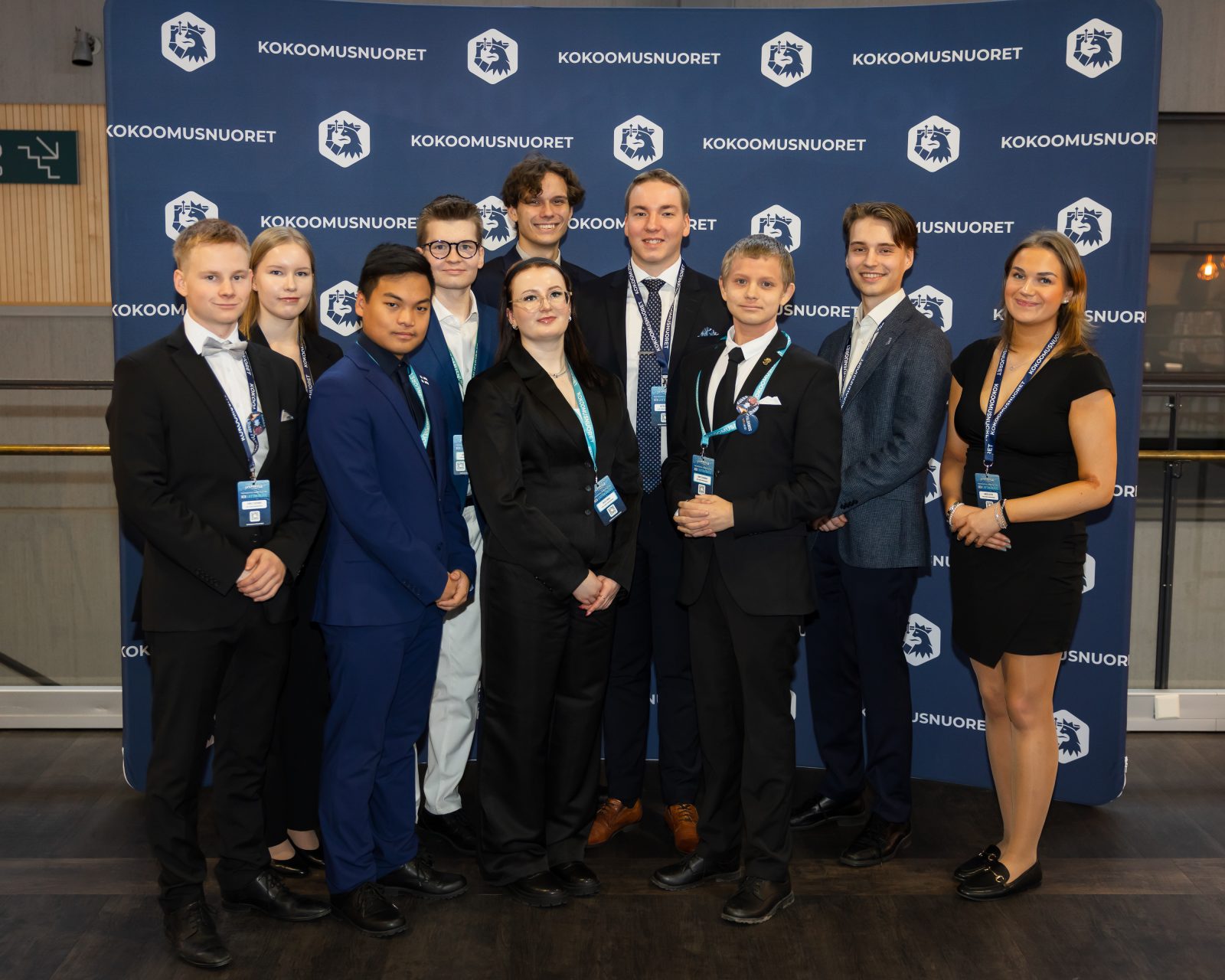 Ryhmäkuva kymmenestä henkilöstä Kokoomusnuorten sinisen kuvausseinän edessä. Kaikki poseeraavat riveissä siisteissä edustus- tai juhlatyylisissä vaatteissa ja katsovat kameraan. Useilla on kaulanauhat ja osallistujapassit kaulassa. Oikeassa reunassa seisova henkilö on pukeutunut mustaan mekkoon, ja eturivissä keskellä on henkilö tummassa puvussa. Taustalla toistuvat Kokoomusnuorten logot.