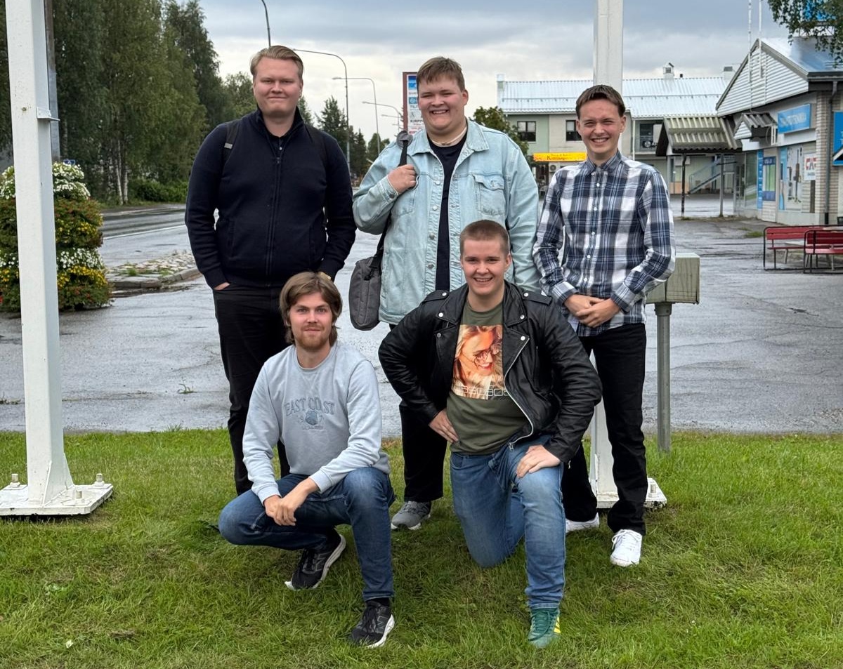 Viisi nuorta aikuista poseeraa ulkona nurmikolla sadepäivänä. Kaksi henkilöä seisoo takarivissä ja kolme on eturivissä, joista yksi kyykistyy nurmelle. Taustalla näkyy märkä katu, matalia rakennuksia, liiketiloja ja puita. Kaikki hymyilevät ja katsovat kohti kameraa.