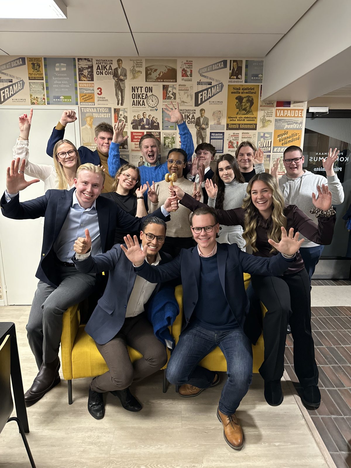 Ryhmäkuva Kokoomusnuorten toimistolta. Etualalla kaksi keltaista nojatuolia, joiden ympärillä ja päällä istuu ja seisoo yhteensä noin viisitoista nuorta aikuista. He poseeraavat iloisesti kädet ylhäällä ja hymyillen kameraan päin. Yksi henkilö pitää kädessään puheenjohtajan nuijaa. Taustalla on seinä täynnä poliittisia julisteita ja Kokoomuksen historiallista kampanjamateriaalia. Kuva on otettu sisätiloissa ja valaistus on kirkas.