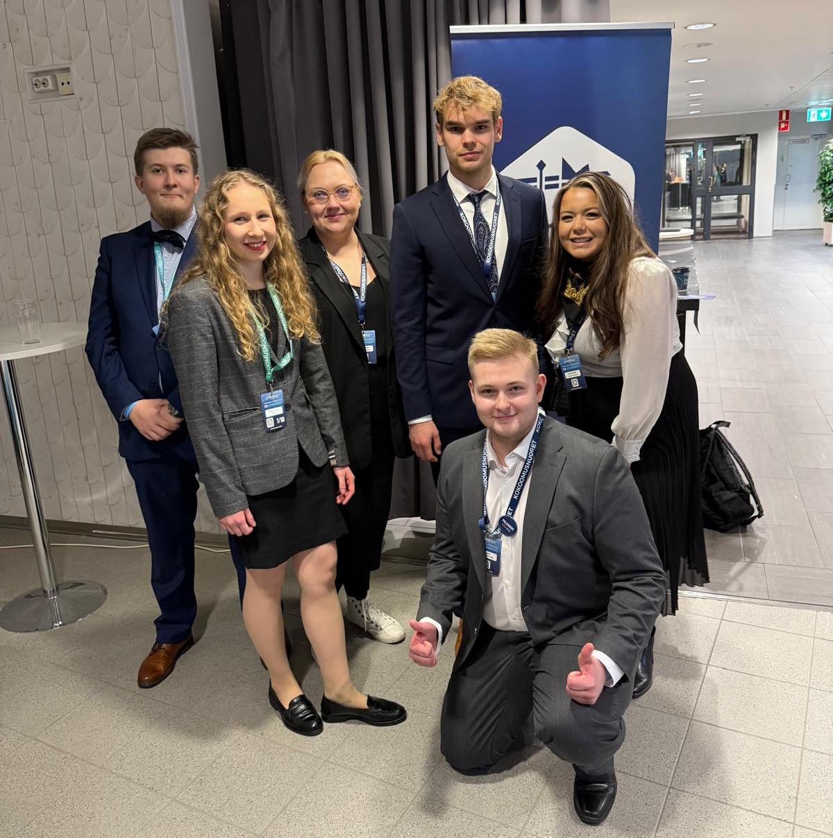 Ryhmäkuva, jossa kuusi nuorta aikuista poseeraa sisätiloissa Kokoomusnuorten tapahtumassa. Taustalla näkyy sininen Kokoomusnuoret-roll-up, harmaa verho ja käytäväalue. Viisi henkilöä seisoo rivissä ja yksi polvistuu etualalle peukut koholla. Kaikilla on osallistujapassit kaulassa, ja he ovat pukeutuneet siististi jakkupuku- tai bisneshenkiseen asuun. Kaikki katsovat kohti kameraa ja hymyilevät.