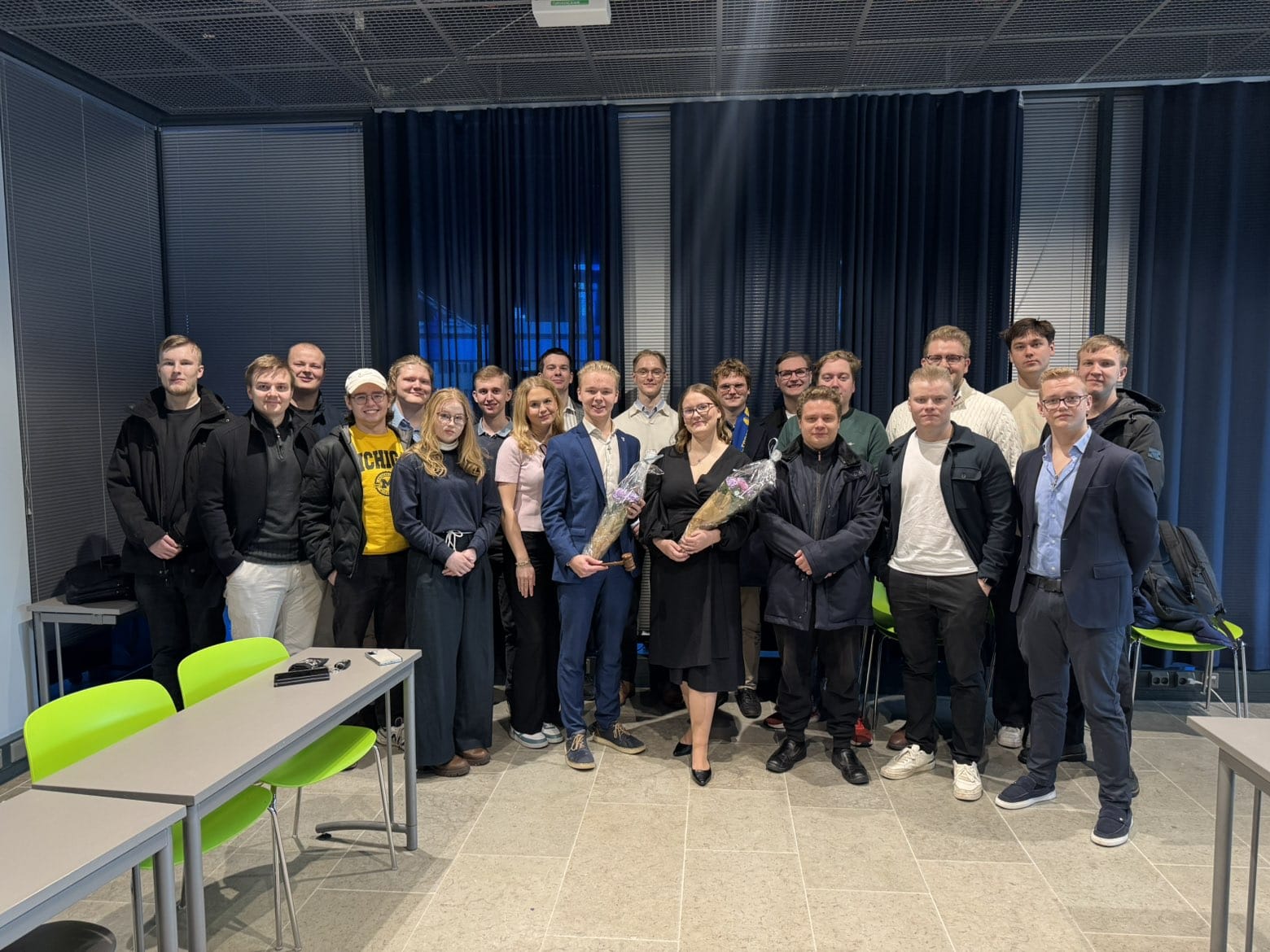 Ryhmäkuva sisätiloissa pidetystä tilaisuudesta. Keskellä kuvaa seisoo noin 20 nuorta aikuista kahdessa rivissä. Eturivissä kaksi henkilöä pitelee kukkakimppuja. Suurin osa ryhmästä hymyilee kameraan ja poseeraa rennosti. Taustalla on tummansiniset verhot ja suuret ikkunat, ja edustalla näkyy harmaita pöytiä sekä vihreitä tuoleja. Kuva on otettu kirkkaassa sisävalaistuksessa.