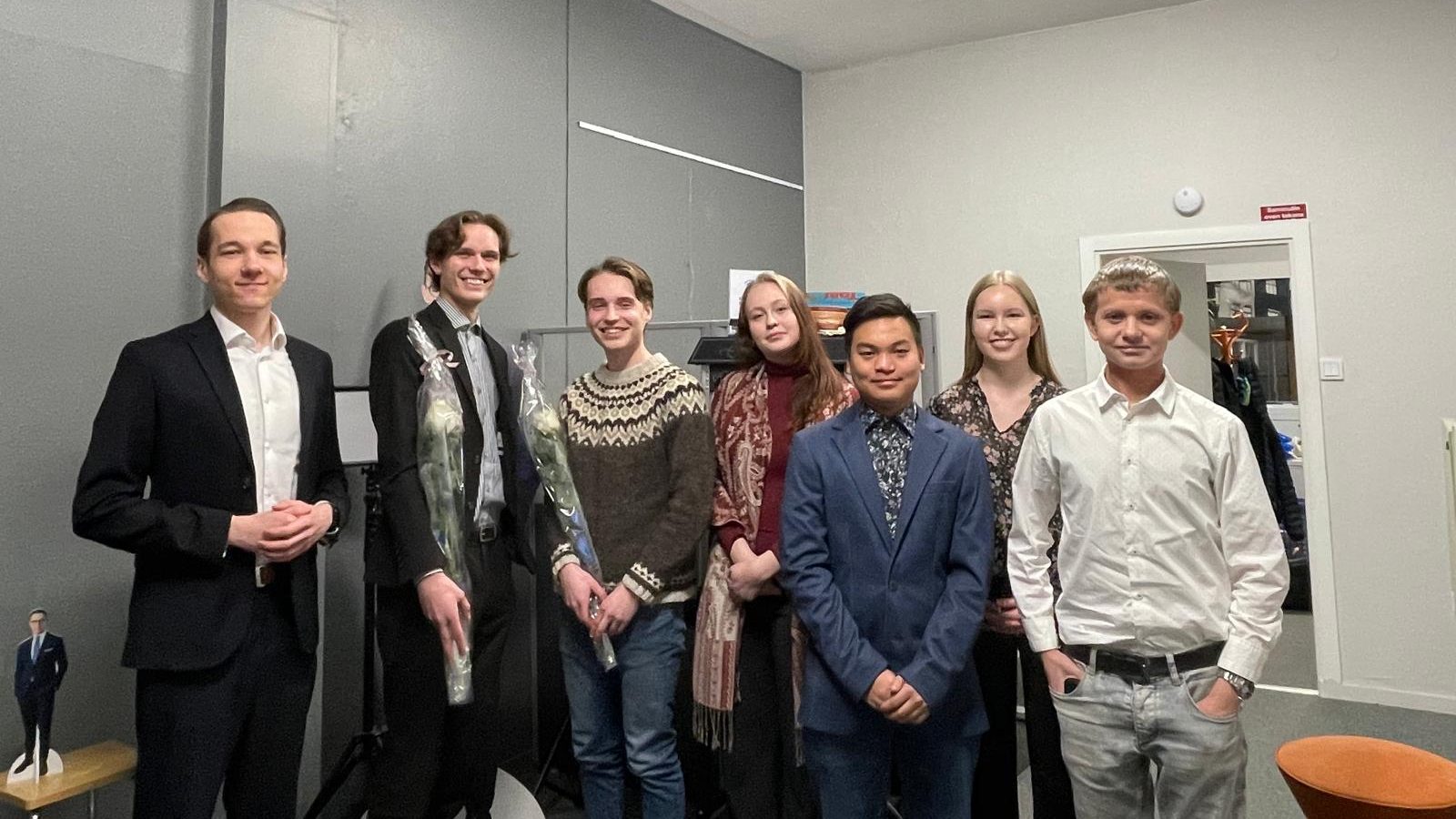 Seitsemän nuorta seisoo rivissä sisätiloissa syyskokouksen jälkeen. Valittu puheenjohtaja ja varapuheenjohtaja pitelevät valkoisia ruusuja. Taustalla näkyy harmaa seinä, ovi ja toimistotilan kalusteita.