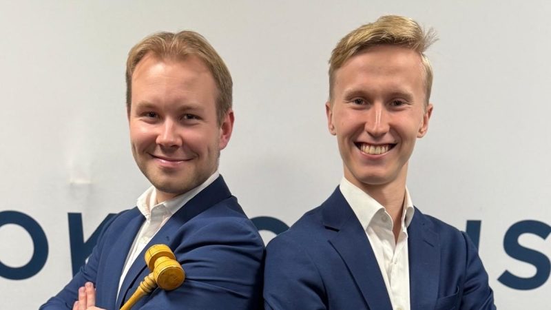 Kaksi nuorta miestä (vasemmalla Jimi Mahonen ja oikealla Juho Virolainen) seisoo selät vastakkain ja hymyilee kameraan. Molemmilla on sininen puku ja valkoinen kauluspaita. Vasemmalla oleva henkilö pitää kädessään puista kokousnuijaa.