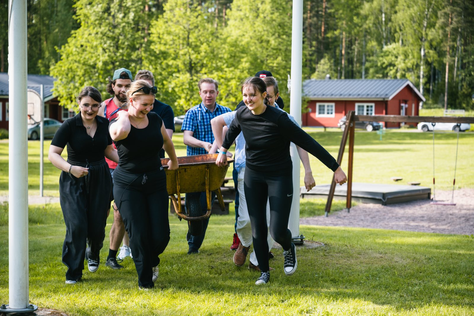 Ryhmä nuoria aikuisia kantaa yhdessä kottikärryjä nurmikolla kesäisessä pihapiirissä. He liikkuvat ryhmänä eteenpäin hymyillen. Taustalla näkyy leikkipaikka, keinut sekä punaisia rakennuksia ja metsäinen ympäristö.