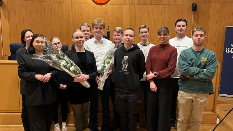 Ryhmäkuva kahdestatoista nuoresta kokoussalissa puupaneloidun seinän edessä. Eturivissä valittu puheenjohtajisto pitelee kukkakimppuja. Taustalla näkyy kokouspöytä ja istuntosalin kalusteita.