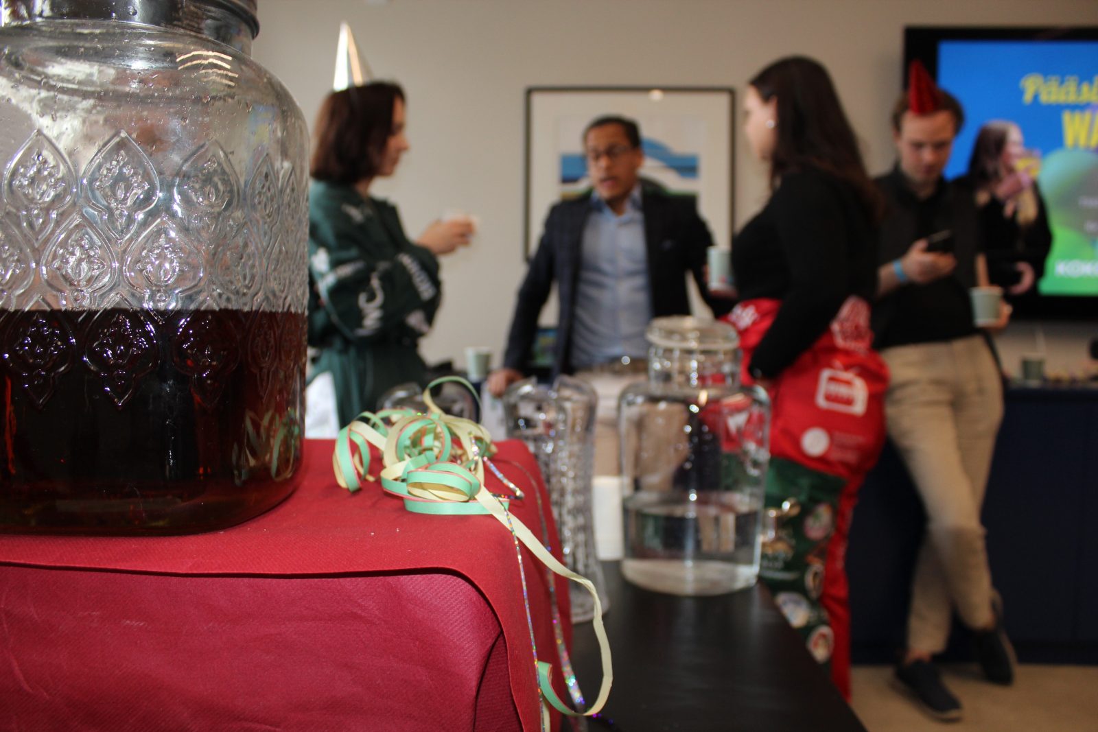 Lasinen booliastia punaisella pöytäliinalla etualalla Kokoomusnuorten pääsihteerin Wappuboolien tarjoilupöydässä. Vieressä on koristeellisia serpentiinejä ja vesikarahveja. Taustalla näkyy ihmisiä keskustelemassa ja juhlimassa sisätilassa vapputunnelmassa.