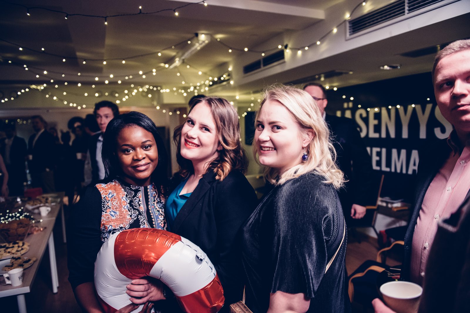 Kolme nuorta naista poseeraa hymyillen kameralle Kokoomusnuorten Sikajuhlilla. Yksi heistä pitää kädessään puna-valkoista karkkitangon muotoista ilmapalloa. Taustalla näkyy juhlavalaistus, tarjoilupöytiä ja muita osallistujia sisätilassa.