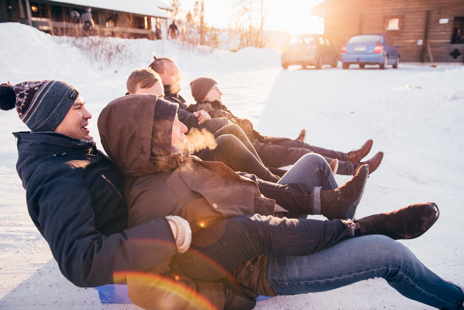 Ryhmä nuoria aikuisia istuu peräkkäin pulkilla lumisella pihalla auringonlaskun aikaan. He ovat pukeutuneet talvivaatteisiin, ja hengitys höyryää kylmässä ilmassa. Taustalla näkyy rakennuksia ja pysäköityjä autoja.