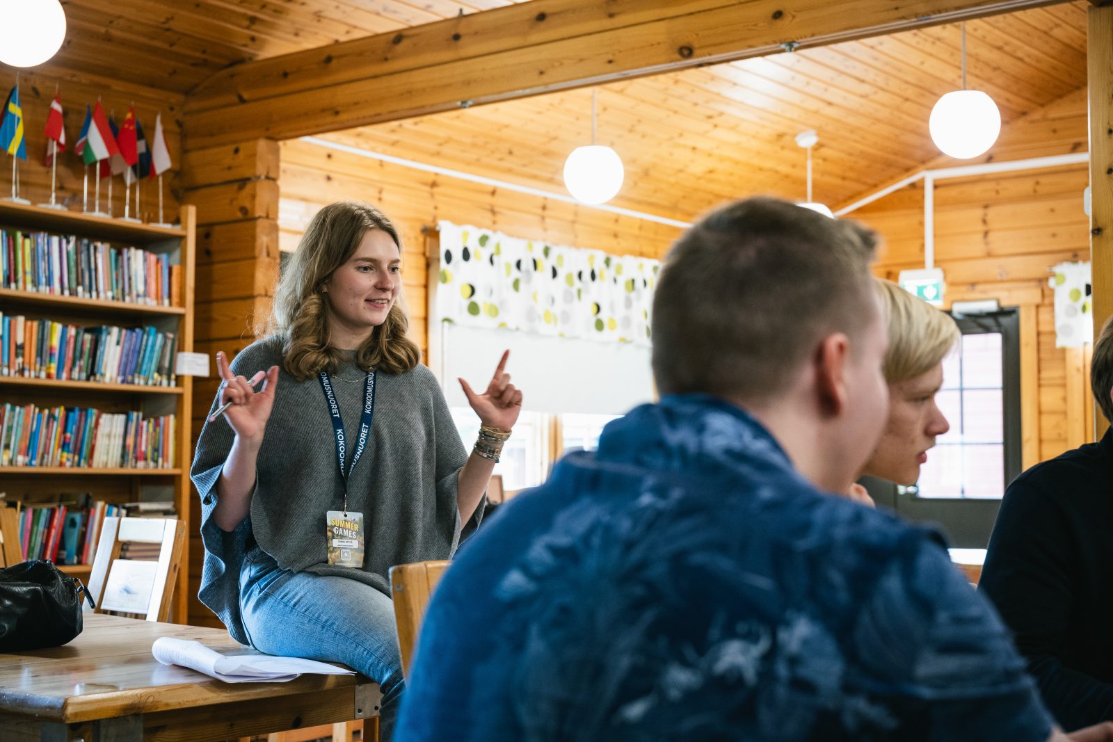 Sisätiloissa otettu kuva puupaneloidussa huoneessa, jossa nainen istuu pöydän reunalla ja puhuu ryhmälle. Hänellä on kaulassaan Kokoomusnuorten tapahtumapassi ja kädet koholla elehtimässä. Etualalla näkyy useita osallistujia selin kameraan. Vasemmalla on kirjahylly täynnä kirjoja ja sen päällä pieniä pöytälippuja. Huoneessa on lämminhenkinen, keskusteleva tunnelma.