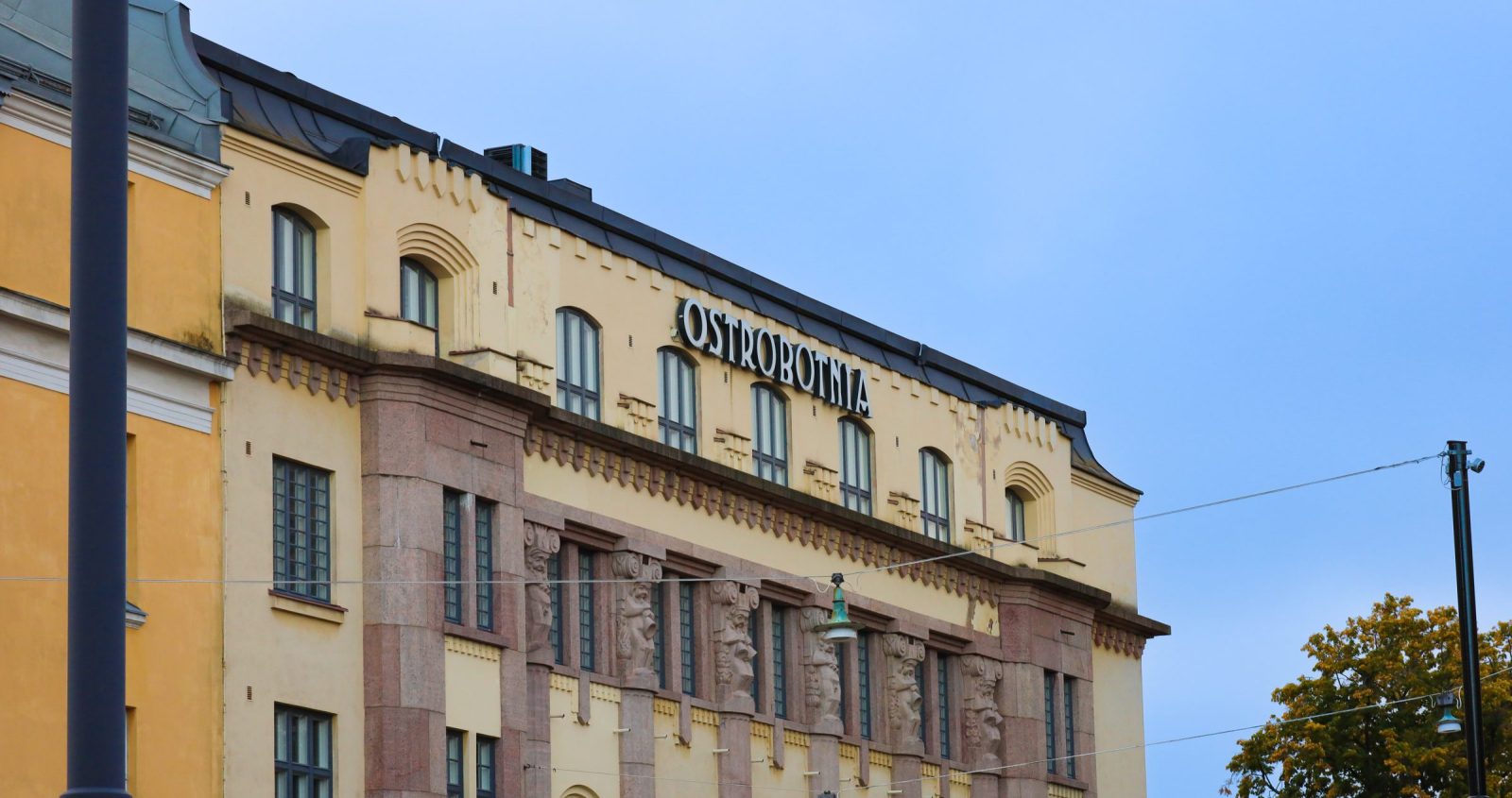 Vaaleankeltainen, koristeellinen kivirakennus, jonka ylimmässä kerroksessa on suuri teksti ”Ostrobotnia”. Julkisivussa näkyy useita pystysuuntaisia ikkunarivejä sekä koristeellisia, vaaleanpunertavasta kivestä veistettyjä yksityiskohtia, kuten ihmishahmoisia reliefipylväitä. Oikeassa reunassa on puu syksyn keltaisine lehtineen ja taustalla vaalea, kirkas taivas.