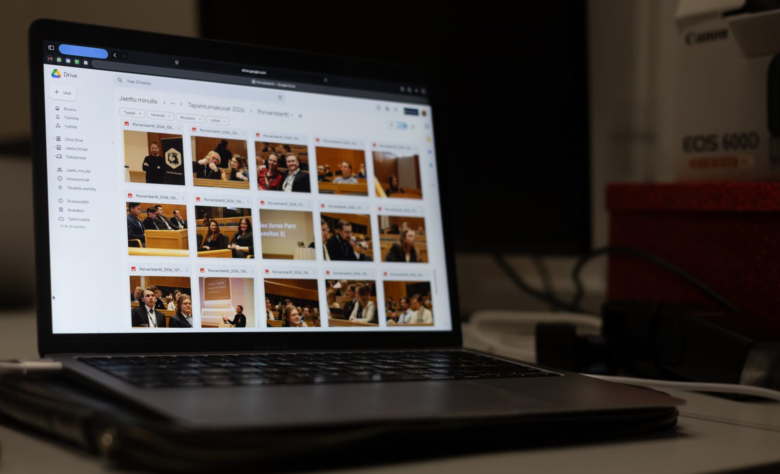Auki oleva kannettava tietokone pöydällä näyttää Google Driven näkymää, jossa on useita pikkukuvia Kokoomusnuorten tapahtumakuvista. Näytön vasemmassa reunassa näkyy Drive-valikko ja oikealla puolella hyllyllä kameratarvikkeita sekä punainen laatikko.