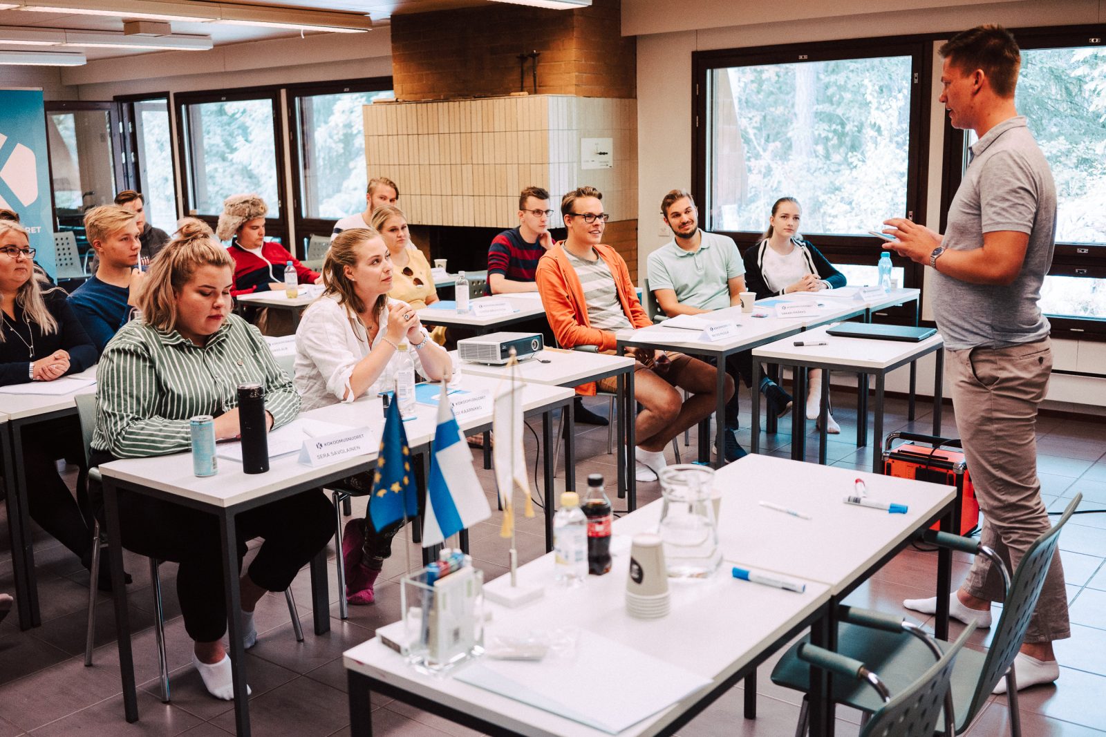 Luokkahuonemaisessa tilassa istuu rivi pöytien ääreen sijoittuneita nuoria aikuisia kuuntelemassa luennoitsijaa, joka seisoo kuvan oikeassa reunassa. Osallistujilla on nimikyltit, muistiinpanovälineitä ja juomapulloja edessään. Etualalla pöydällä näkyy Suomen ja Euroopan unionin pienet pöytäliput sekä kannu ja kertakäyttömukeja. Taustalla suuret ikkunat avautuvat metsänäkymään. Tunnelma on keskittynyt ja opintotyöhön paneutunut.