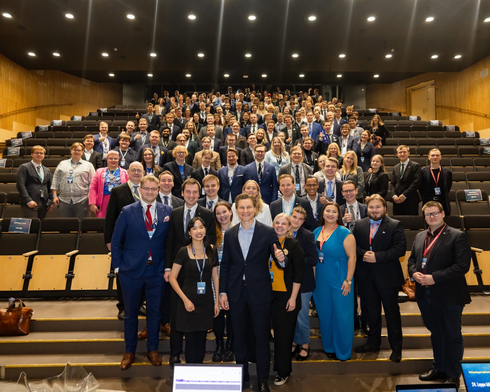 Suuri joukko Kokoomusnuorten liittokokouksen osallistujia poseeraa auditoriossa. Etualalla seisoo useita nuoria aikuisia hymyillen ja katsoen kameraan, osa näyttää peukkua. Taustalla näkyy kymmenittäin muita osallistujia istumapaikoilla ja portailla, kaikki pukeutuneina siisteihin kokous- tai juhla-asuihin. Valoisa auditorio on täynnä ihmisistä muodostuvaa ryhmäkuviota.