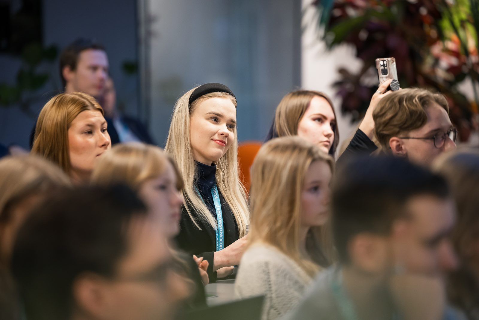 Yleisö istuu sisätiloissa seuraamassa esitystä tai puhetta. Etualalla näkyy useita nuoria aikuisia, joista keskellä istuva vaaleahiuksinen nainen hymyilee kevyesti ja katsoo eteenpäin. Hänellä on tummat vaatteet ja Kokoomusnuorten tapahtuman osallistujapassi kaulassa. Vieressä ja ympärillä muut osallistujat kuuntelevat tarkkaavaisina; yksi henkilö takana nostaa puhelimen ylös ottaakseen kuvan tai videon. Taustalla näkyy kasveja ja epäterävästi rajautuneita osallistujia.