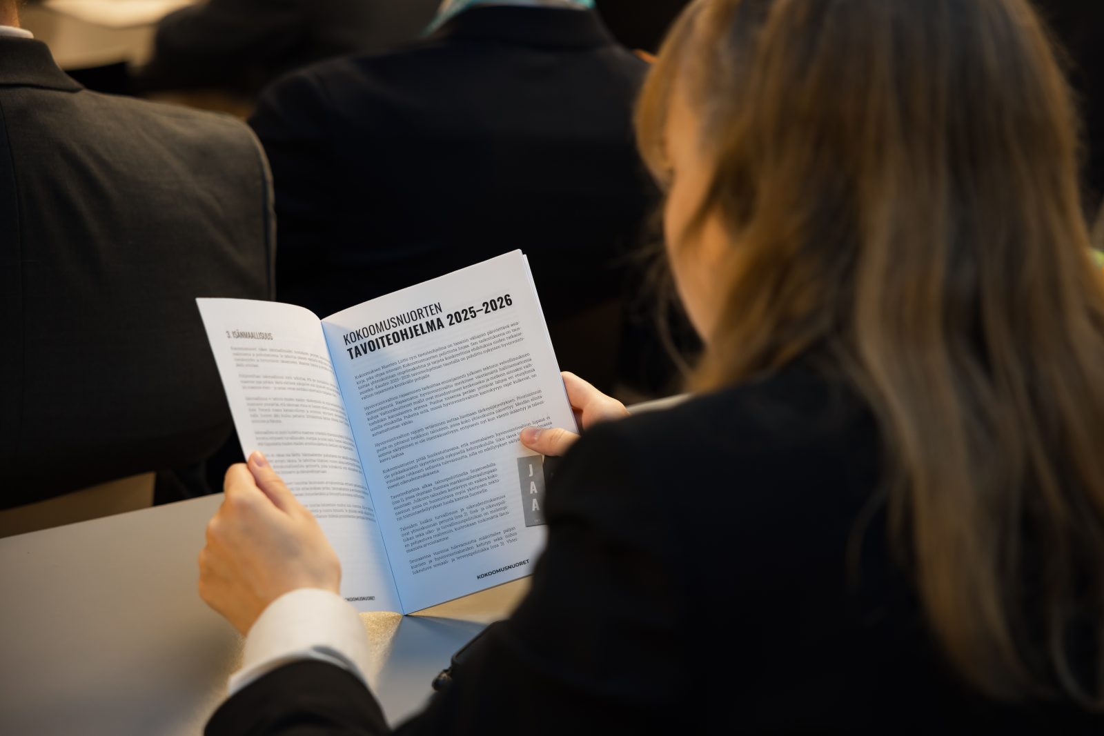 Lähikuva takaapäin nuoresta naisesta, joka lukee käsissään olevaa vihkoa. Vihon oikealla sivulla on otsikko ”Kokoomusnuorten tavoiteohjelma 2025–2026”. Nainen istuu yleisössä, ja hänen edessään näkyy muiden osallistujien selkiä tummissa puvuissa. Kuva on otettu Porvaristartti 2025 -tapahtumassa.