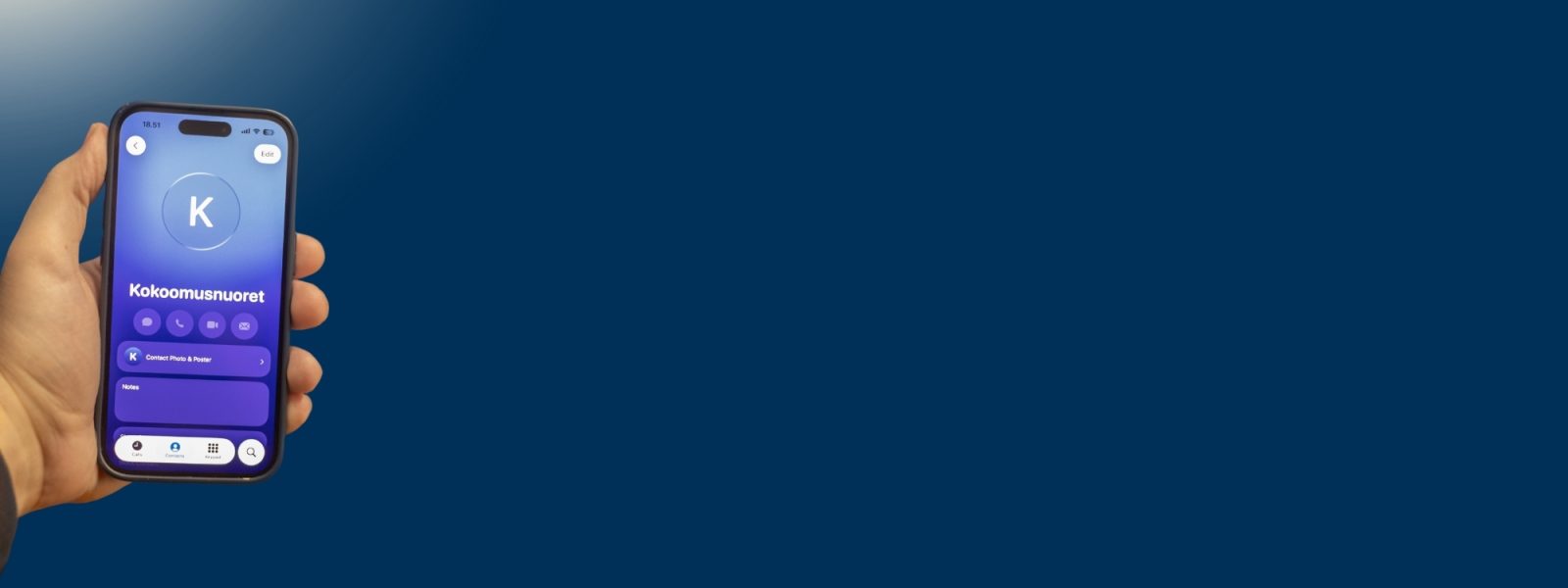 Käsi pitää älypuhelinta tummansinistä seinää vasten. Puhelimen näytöllä näkyy yhteystietokortti nimellä ”Kokoomusnuoret”, sinisävyisellä taustalla ja useilla pikapainikkeilla (soitto, viesti, sähköposti). Tausta on yksivärinen ja tyhjä, mikä korostaa puhelimen näyttöä.