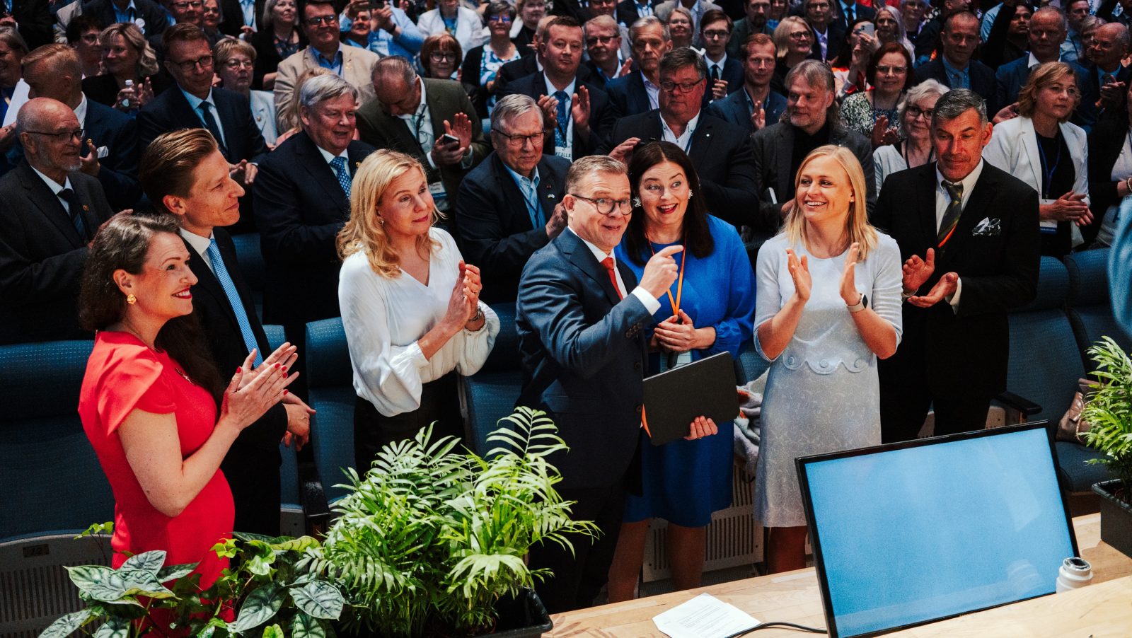 Kokoomuksen johtoon kuuluvia ihmisiä seisoo puoluekokouksessa suuressa salissa. Etualalla on poliitikkoja, jotka taputtavat ja hymyilevät katsomoon päin; Petteri Orpo osoittaa jotakin ja pitää mapin kädessään. Taustalla yleisö seuraa tilannetta ja aplodeeraa. Tunnelma on iloinen ja virallinen, ja kuva on täynnä ihmisiä.