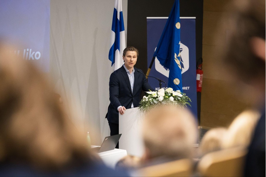 Antti Häkkänen seisoo puhujakorokkeen takana ja pitää puhetta kokoussalissa. Korokkeen edessä on kukkalaite. Taustalla näkyy Suomen lippu sekä Kokoomusnuorten sininen lippu ja tapahtumabanneri. Etualalla näkyy epäselvästi yleisön jäsenten päitä ja olkapäitä.