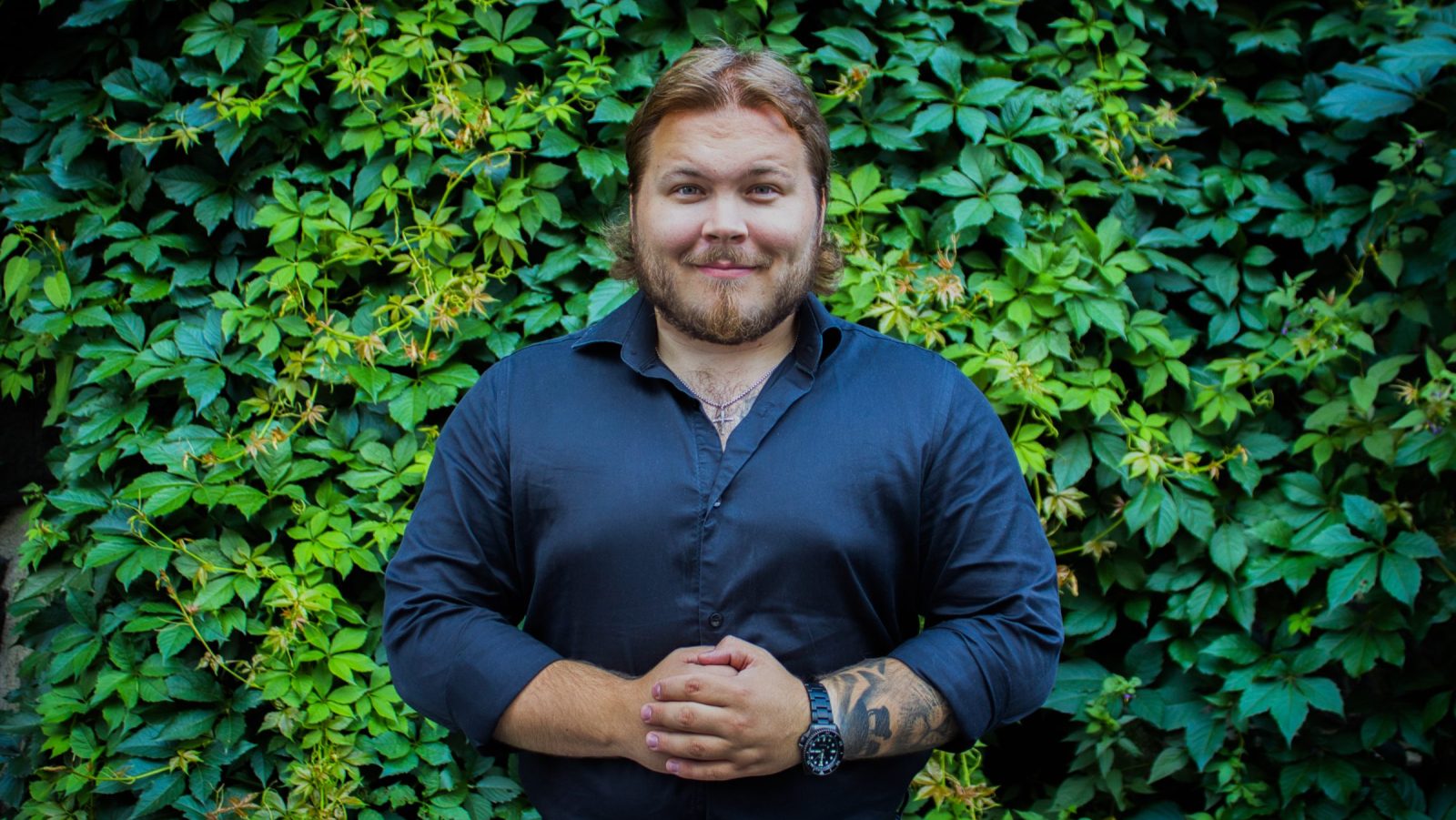 Kuvassa mies seisoo vihreän köynnösseinän edessä ja katsoo kameraan. Hänellä on päällään musta kauluspaita, ja hänen kätensä ovat kevyesti yhdessä edessä. Hänellä on rannekello ja tatuointi käsivarressa, ja hän hymyilee.