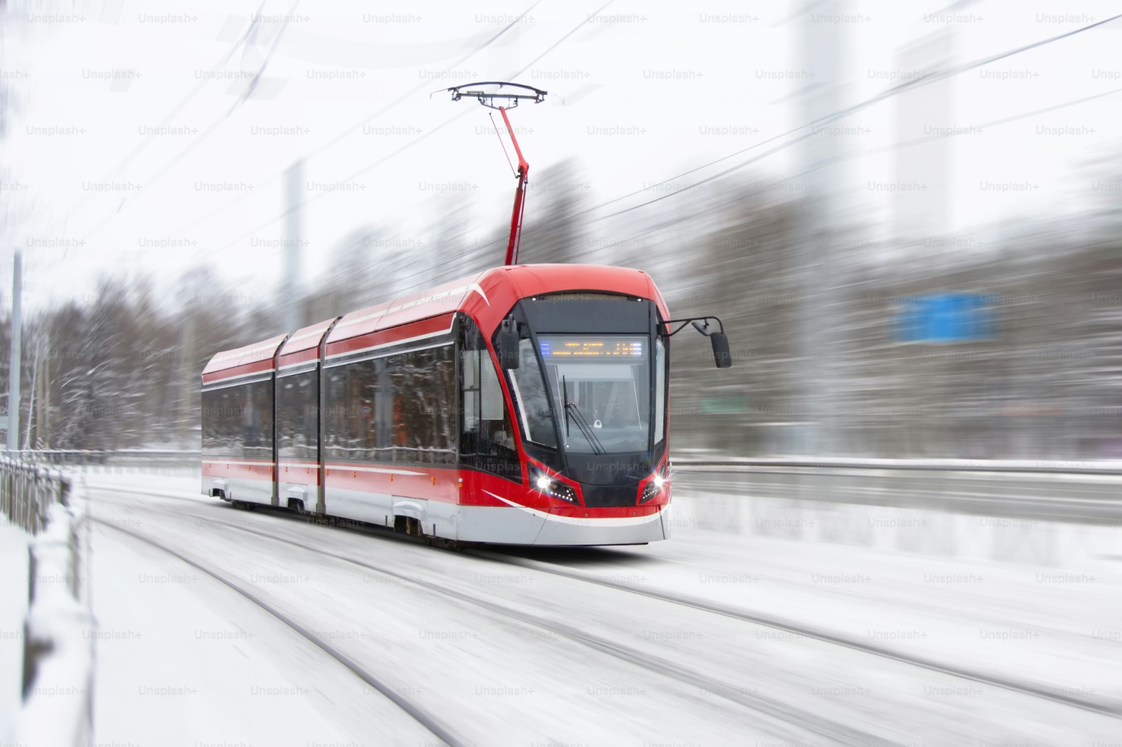 Valokuva modernista punaisesta raitiovaunusta, joka kulkee lumisessa kaupunkiympäristössä. Liike-epäterävyys korostaa vauhtia. Tausta on talvinen ja vaalea. Kuvassa on Unsplash-kuvapalvelun vesileima.