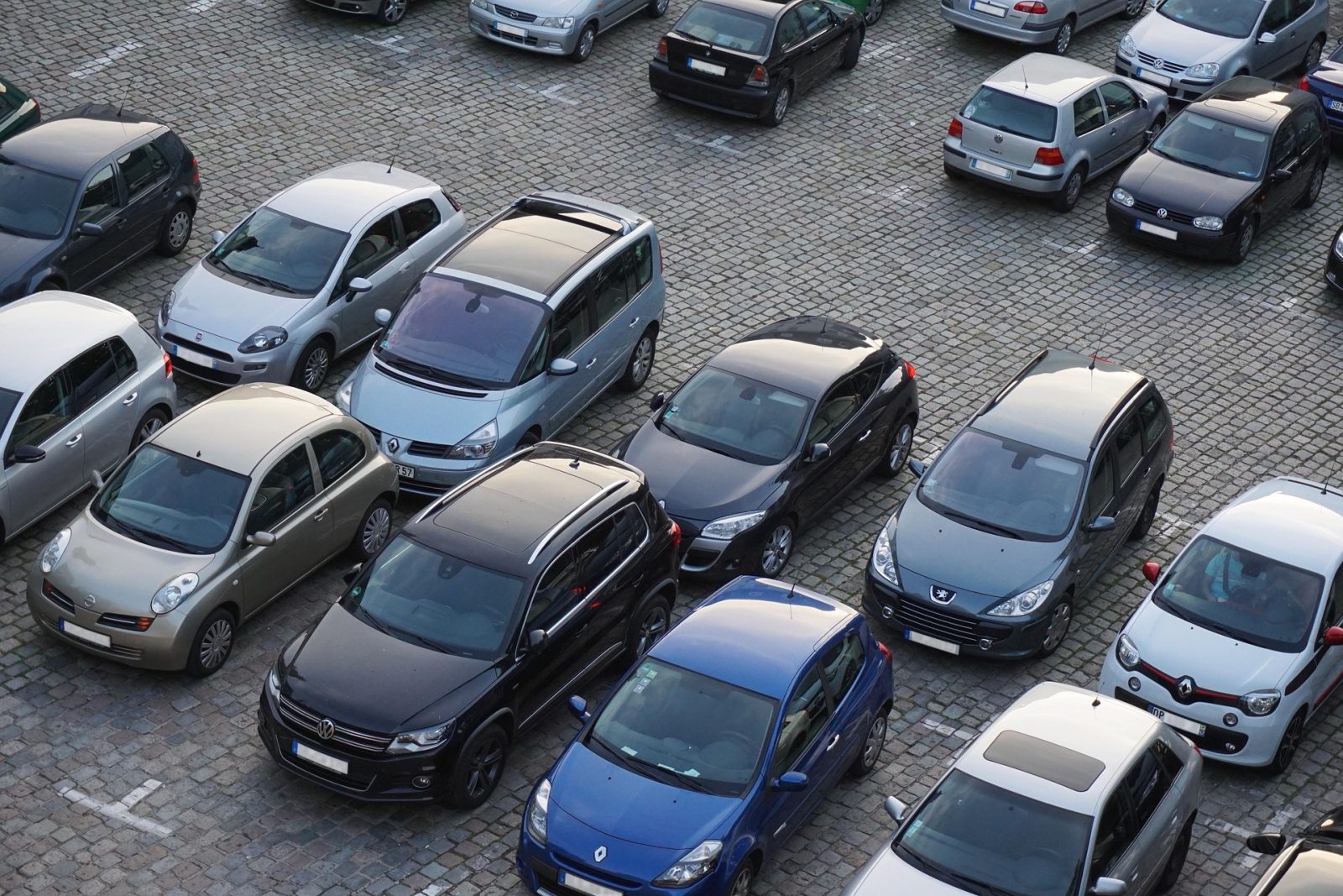 Valokuva täynnä olevasta parkkipaikasta ylhäältä kuvattuna. Kuvan täyttävät erikokoiset ja -väriset henkilöautot, jotka on pysäköity epäjärjestyksessä kivipaatein päällystetylle alueelle.