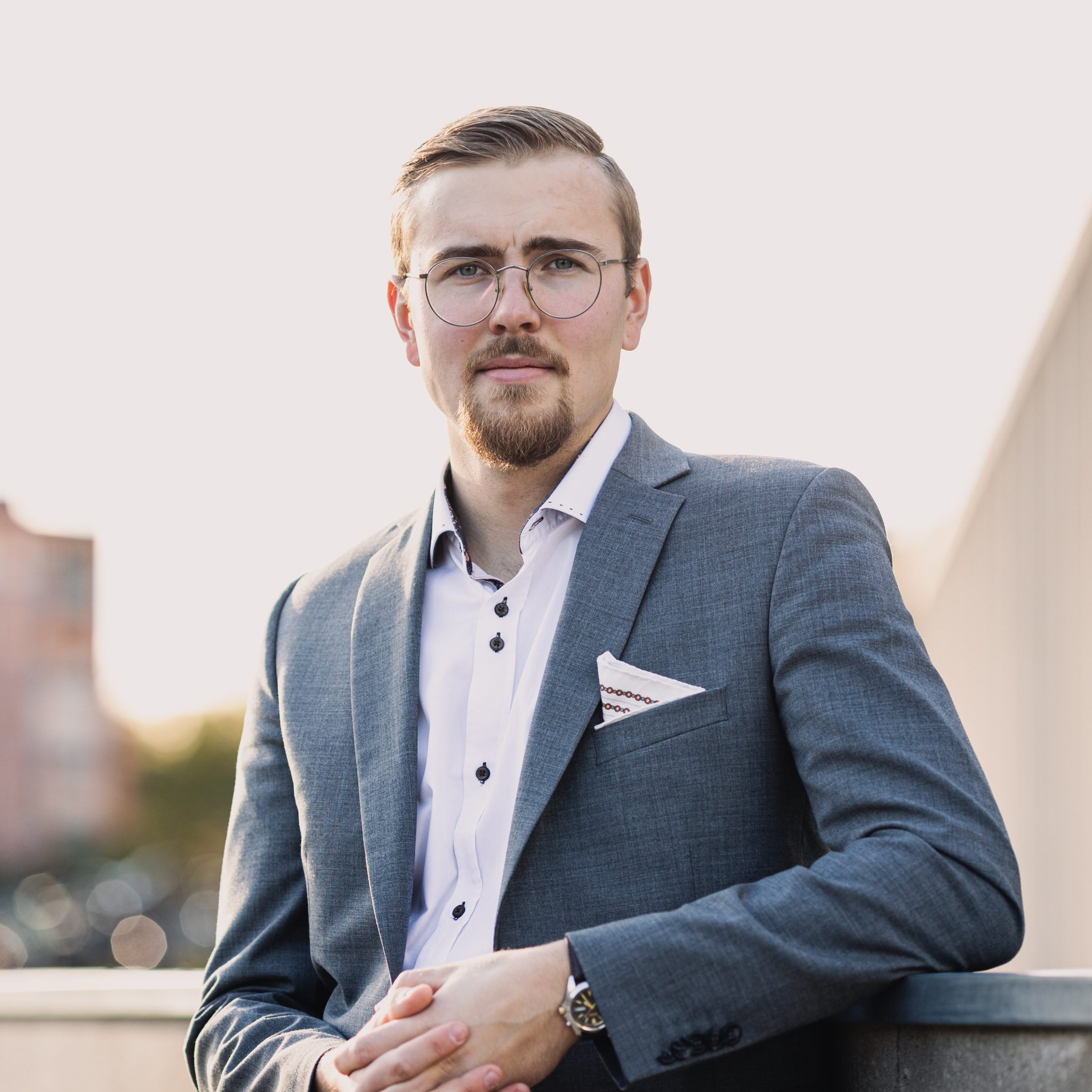Muotokuvavalokuva Joona Paijasta harmaassa bleiserissä, valkoisessa kauluspaidassa ja pyöreissä silmälaseissa nojaamassa kaiteeseen. Taustalla näkyy epäterävä kaupunkimaisema.