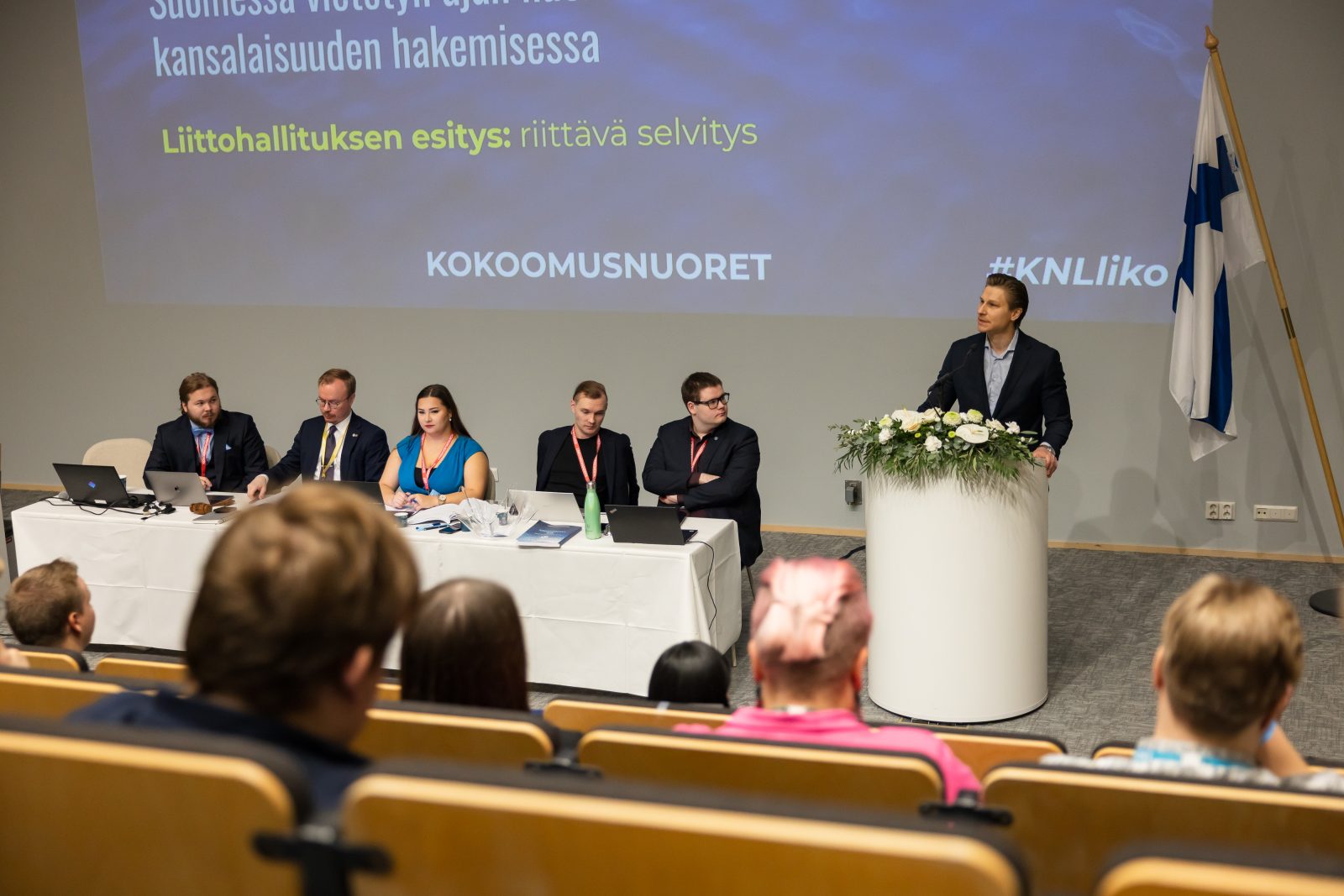 Valokuva Kokoomusnuorten liittokokouksesta. Etualalla istuu yleisöä, taustalla presidiumin pöydässä istuu viisi henkilöä kannettavien tietokoneiden ääressä. Oikealla puhujanpöntöllä seisoo mies tummassa puvussa, vieressä Suomen lippu. 