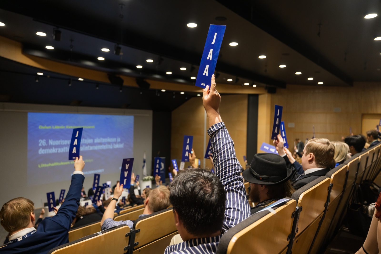 Valokuva Kokoomusnuorten liittokokouksesta, jossa äänestäjät nostavat sinisiä "Jaa"-äänestyslappuja ilmaan kokousauditoriossa. Taustalla näkyy diaesitys, jossa on käsiteltävänä oleva aloite.