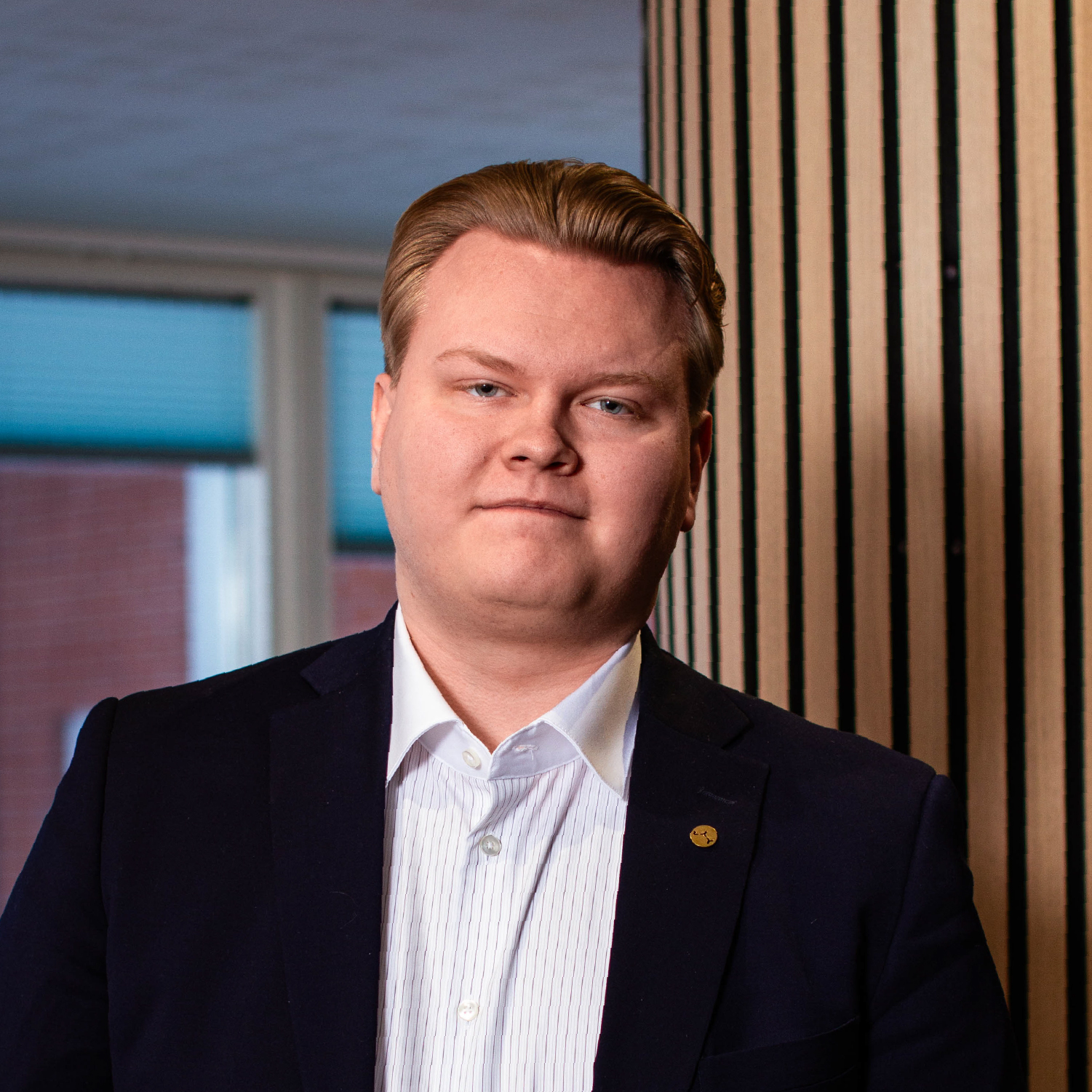 Muotokuvavalokuva Timo Korkiakoskesta tummassa puvussa ja raidallisessa kauluspaidassa kultainen rintamerkki siinpelissä. Hän katsoo suoraan kameraan neutraalilla ilmeellä. Taustalla näkyy puuverhottua seinää ja ikkunoita.
